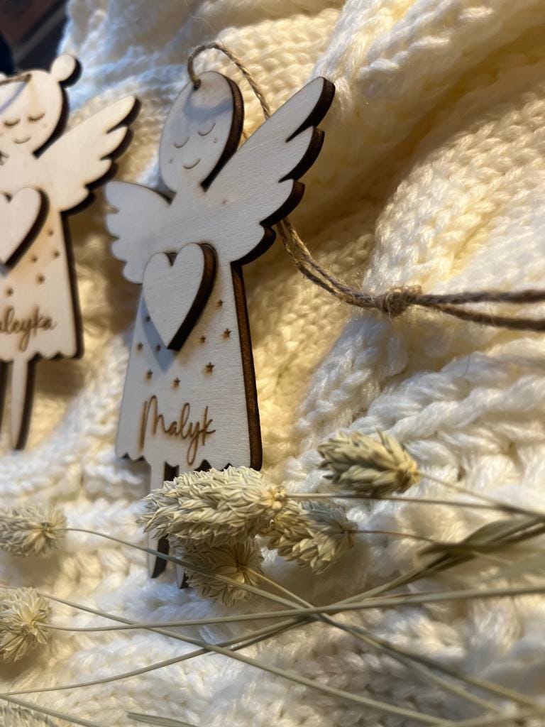 Personalisierter Schutzengel aus Holz mit Namen Malyk, aufgehängt an einer Kordel vor heller Strickdecke